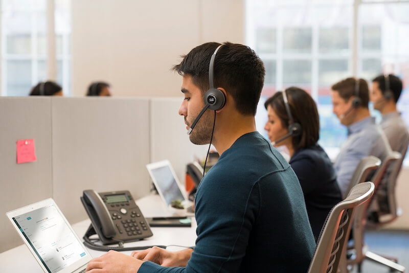 EIn Mann telefoniert im Großraumbüro über das Headset und das Cisco-Telefon, während er etwas in den Laptop einträgt
